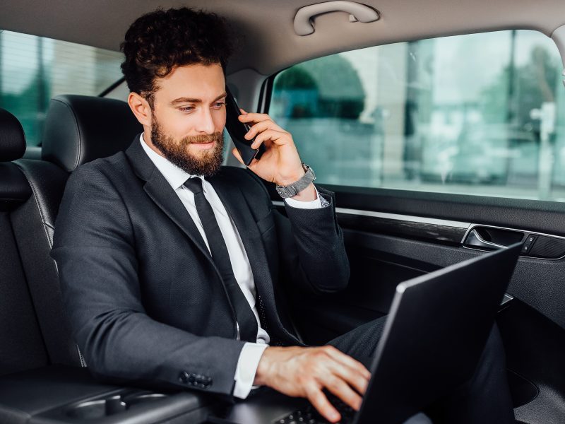 Handsome, bearded, smiling businessman working on his laptopand speaking mobile phone on the backseat of the car.